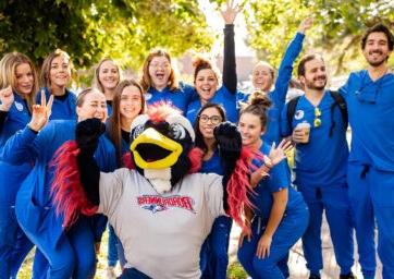 Healthcare students in royal blue scrubs posing for a picture with Rowdy on a sunny, fall day